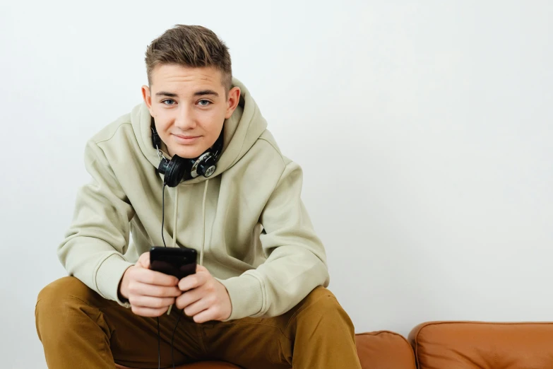 a man sitting on a couch holding a cell phone, hyperrealistic teen, wearing black headphones, promo image, looking at camera