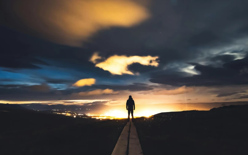 a person standing on top of a wooden walkway, by Niko Henrichon, pexels contest winner, well lit sky, dramatic backgroung, in the distance, back lit