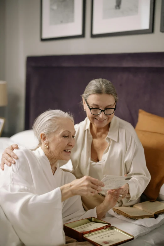 a couple of women sitting on top of a bed, gray haired, checking her phone, promo image, multiple stories