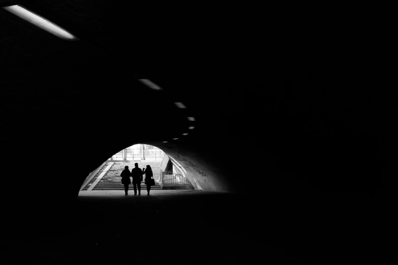 a couple of people that are standing in a tunnel, a black and white photo, by Cedric Peyravernay, unsplash contest winner, minimalism, three women, traversing a shadowy city, underlit, hunters