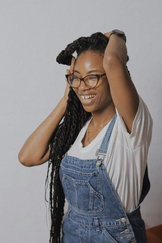 a woman with glasses standing in front of a white wall, by Lily Delissa Joseph, wearing blue jean overalls, long dreadlocks, smiling playfully, hands in her hair