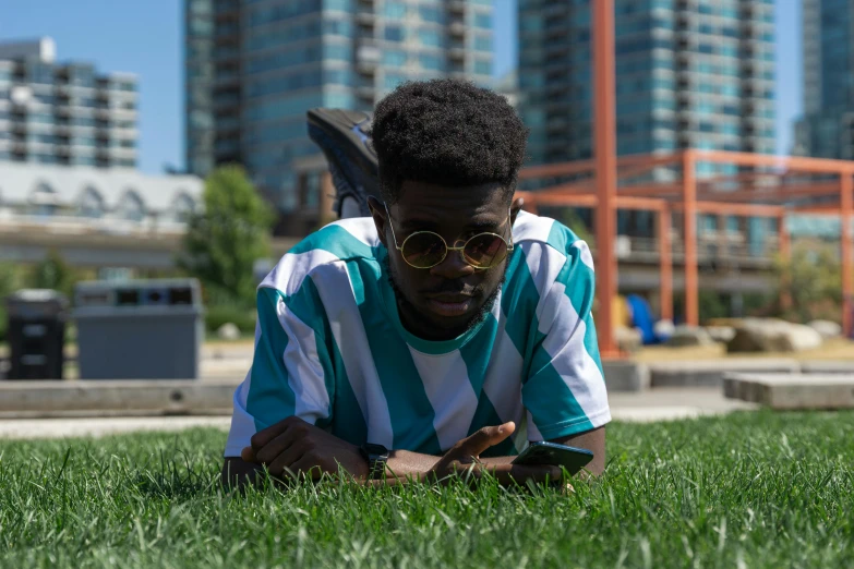 a young man laying on top of a lush green field, by Andrew Stevovich, pexels contest winner, realism, afro tech, with teal clothes, in the city, toronto