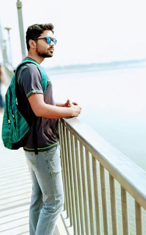 a man standing on a bridge next to a body of water, a picture, inspired by Bikash Bhattacharjee, with a backpack, wearing green, posed in profile, overexposed photograph