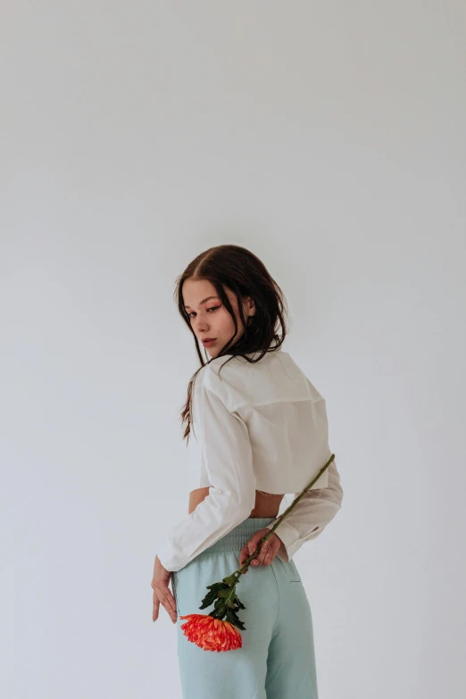 a woman standing in front of a white wall holding a flower, an album cover, by Emma Andijewska, trending on unsplash, renaissance, white shirt and green skirt, portrait sophie mudd, lower back, wearing a light blue shirt