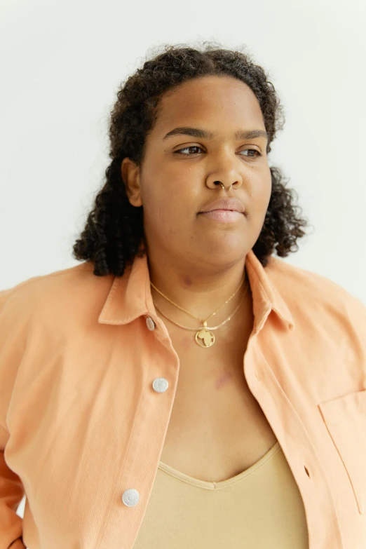 a woman standing with her hands in her pockets, by Winona Nelson, wearing gold detailed choker, lizzo, wear's beige shirt, wearing several pendants