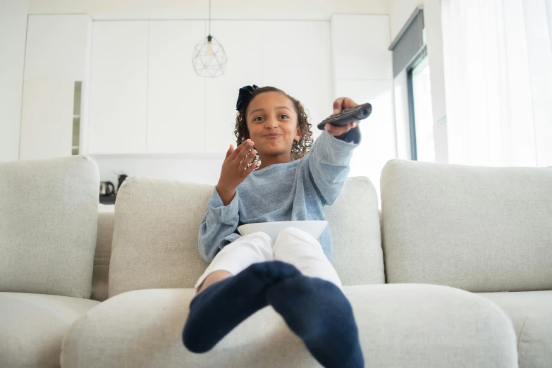 a little girl sitting on a couch holding a remote control, pexels, happening, girl wearing uniform, light skinned african young girl, 1 2 9 7, multiple stories