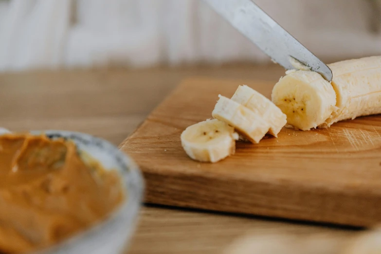 a banana sitting on top of a cutting board next to a bowl of peanut butter, by Sylvia Wishart, trending on pexels, holds a small knife in hand, a wooden, manuka, 🦩🪐🐞👩🏻🦳