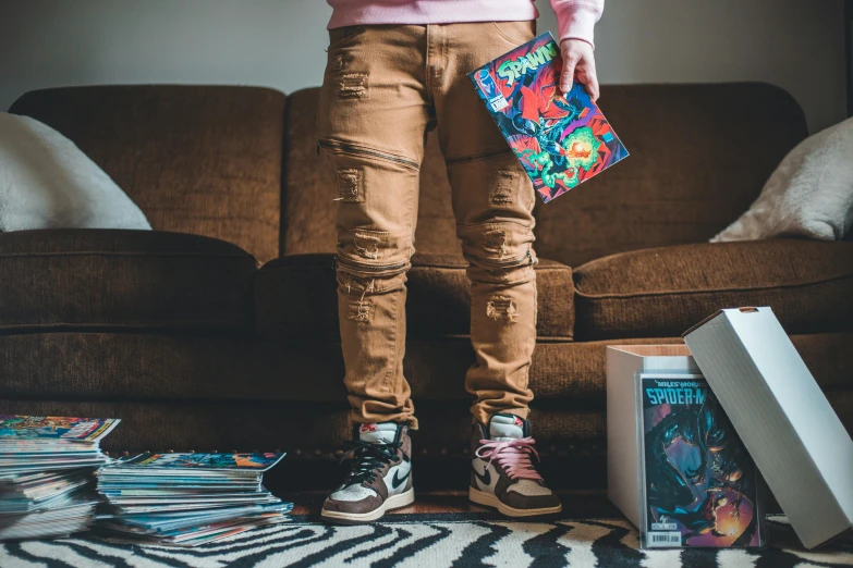 a person standing in front of a couch holding a book, an album cover, inspired by NEVERCREW, trending on pexels, graffiti, ripped jeans, colorful comics style, sneaker photo, brown pants