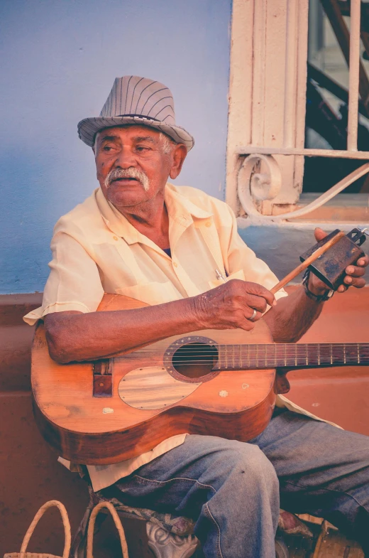 a man with a hat on sits in front of a window playing a guitar