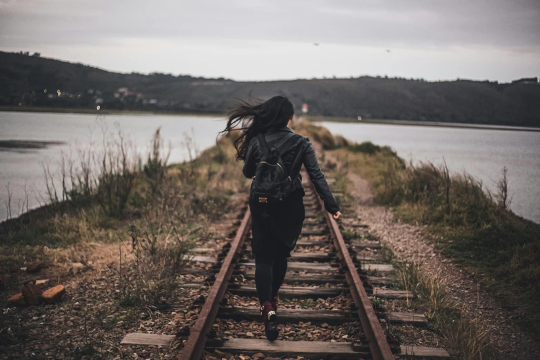 a woman walking down a train track next to a body of water, pexels contest winner, grungy woman, 🚿🗝📝, with a backpack, female model