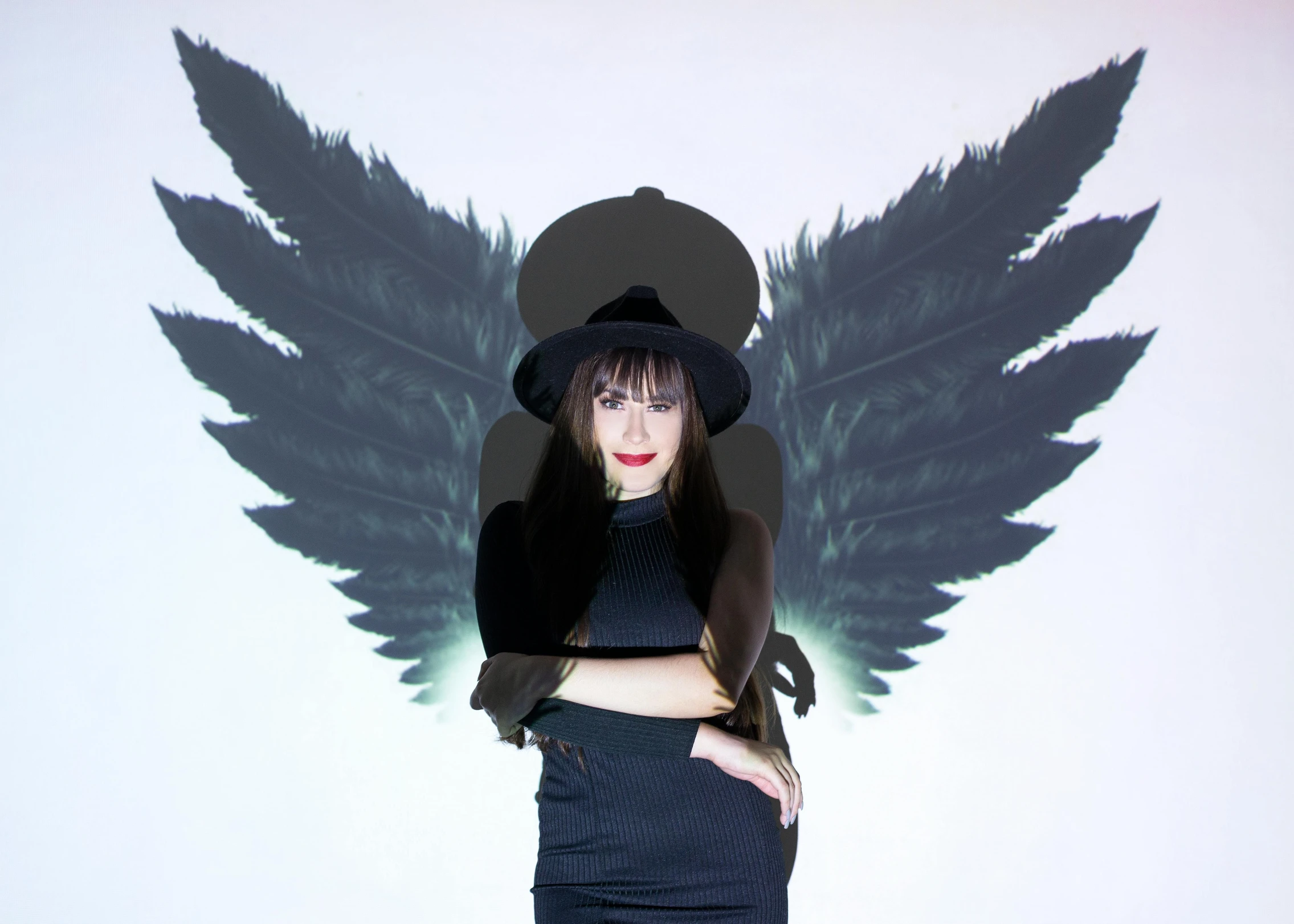 a woman standing in front of a wall with wings on it, an album cover, inspired by Julia Pishtar, wearing black witch hat, cardboard cutout, charli bowater, ( ( ( black witch hat ) ) ) )