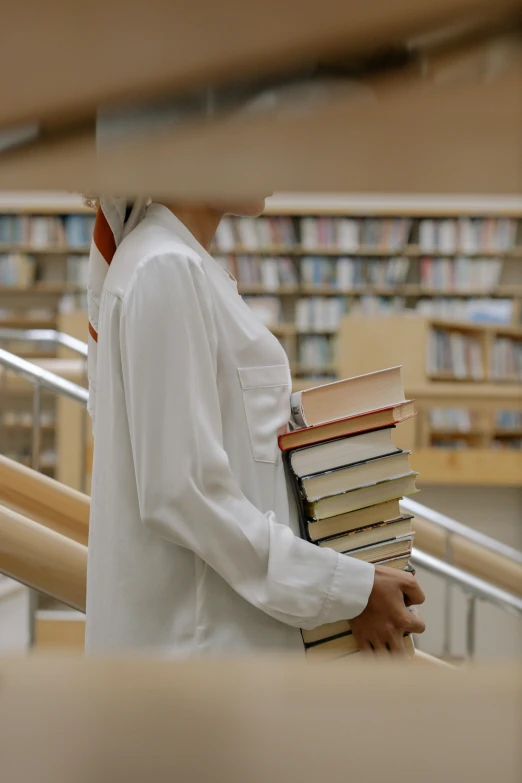 a man holding a stack of books in a library, an album cover, by Matija Jama, unsplash, wearing lab coat and a blouse, white hijab, walking to the right, low quality footage