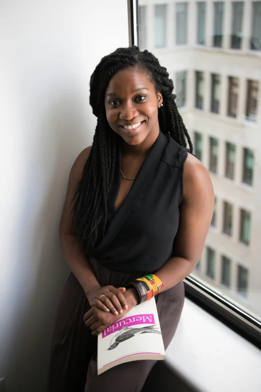 a woman standing in front of a window holding a book, by Lily Delissa Joseph, posing with crossed arms, dark skinned, vp of marketing, adafruit