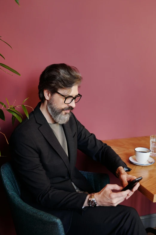 a man sitting at a table using a cell phone, with a beard and a black jacket, wearing black frame glasses, ignant, luka mivsek