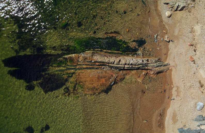 a boat sitting on top of a sandy beach next to a body of water, a digital rendering, by Attila Meszlenyi, land art, geological strata, looking down on the camera, sea serpent, photo from the dig site