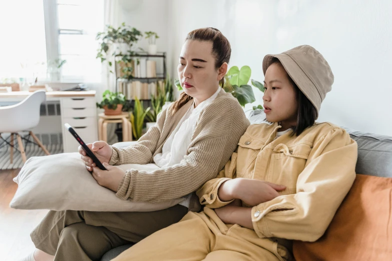 two women sitting on a couch looking at a cell phone, a cartoon, trending on pexels, asian decent, avatar image, lonely family, wearing a yellow hoodie