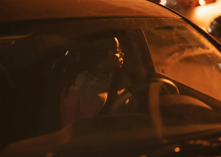 a woman sitting in the driver's seat of a car, by Dan Frazier, pexels contest winner, dark-skinned, ambient amber light, reflecting, low quality footage