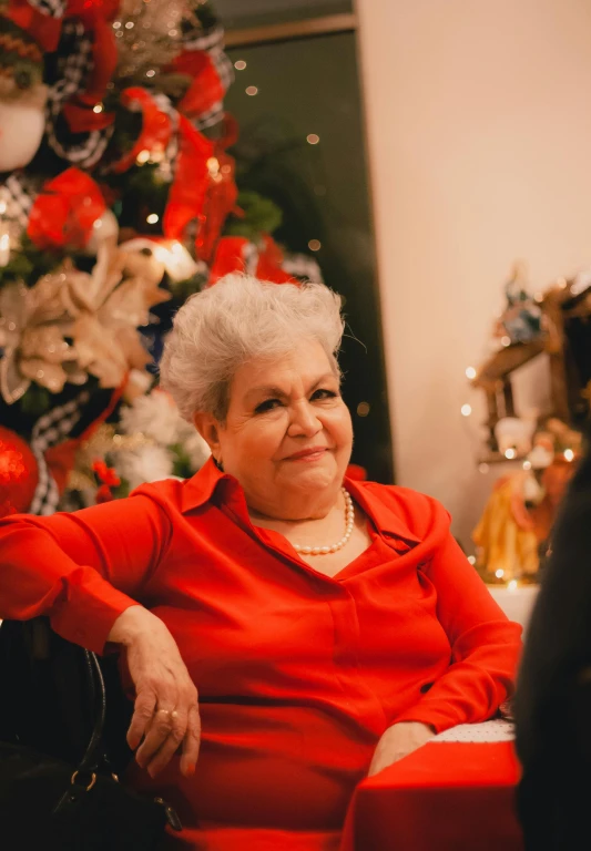 an elderly woman in a red dress sits under a christmas tree