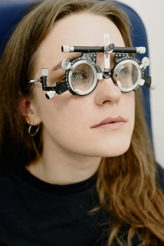 a woman sitting in a chair with a pair of glasses on her face, by Adam Marczyński, trending on unsplash, photorealism, metal eye piece, white prosthetic eyes, portrait of a female pathologist, stars as eyes