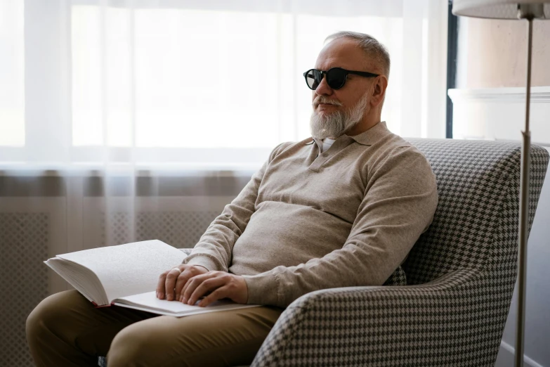 a man sitting in a chair reading a book, wearing shades, grey beard, sitting across the room, profile image