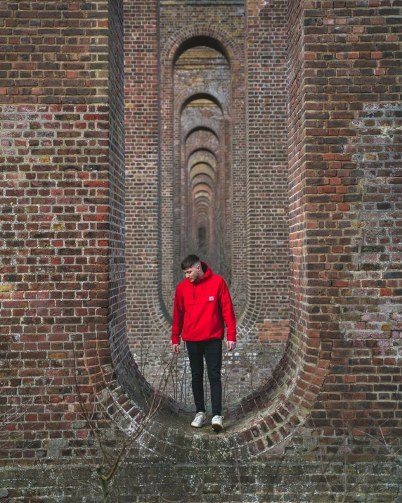 a man in a red jacket standing in front of a brick wall, inspired by Storm Thorgerson, pexels contest winner, high walled tunnel, view(full body + zoomed out), crystal palace, structure : kyle lambert