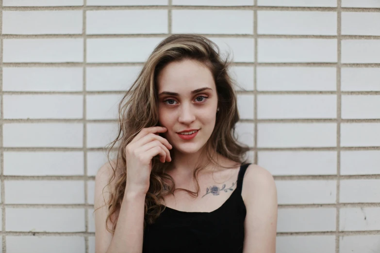 a woman standing in front of a white brick wall, inspired by Elsa Bleda, pexels contest winner, photorealism, with long hair and piercing eyes, she is wearing a black tank top, natalia dyer, pale skin!