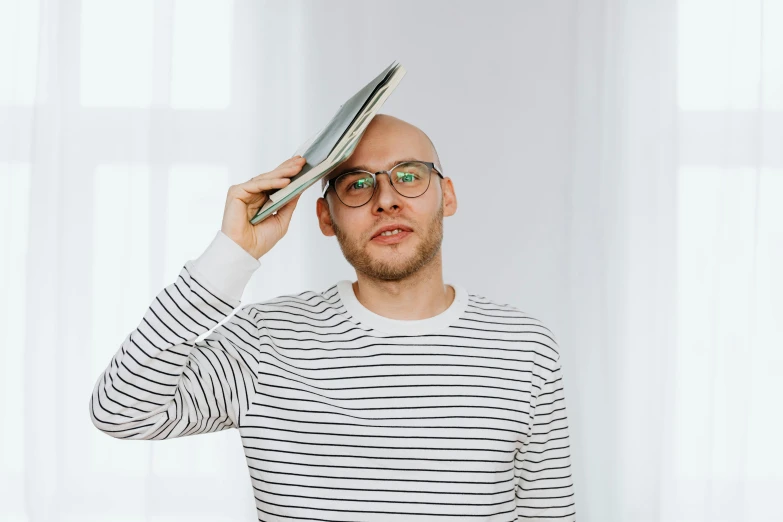 a man in a striped shirt holding a book over his head, an album cover, by Julia Pishtar, pexels contest winner, bald patch, rounded eyeglasses, andrey surnov, looking to the side