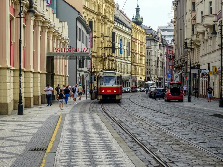 a red train traveling down a street next to tall buildings, by karolis strautniekas, pexels contest winner, art nouveau, in legnica, trams ) ) ), square, cobblestone road