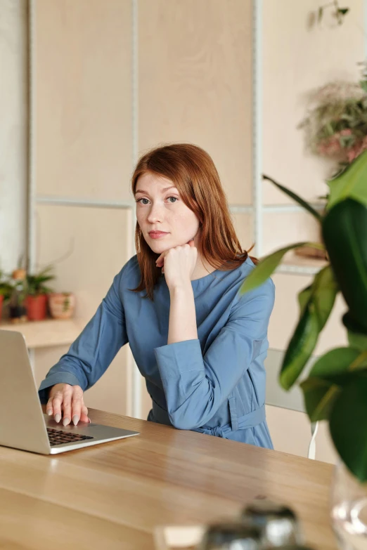 a woman sitting at a table with a laptop, next to a plant, wearing a light blue shirt, redhead woman, thoughtful pose