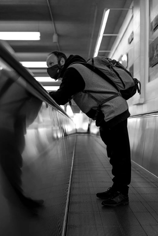 a black and white photo of a man on an escalator, wearing a mask, backpack, face down, underground