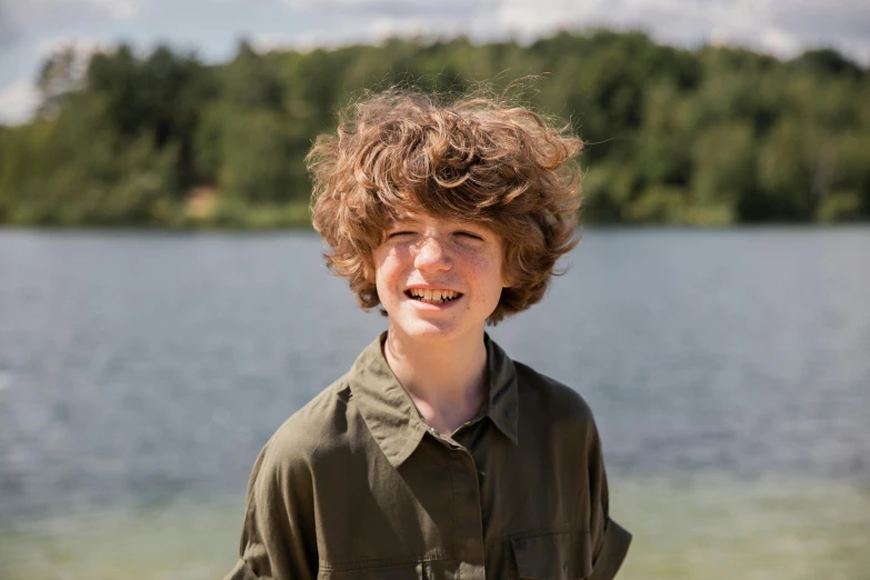 a young boy standing in front of a body of water, ginger hair with freckles, while smiling for a photograph, lakeside, bartlomiej gawel