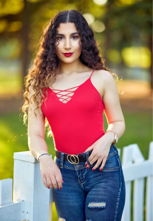 a woman standing next to a white fence, wearing red tank top, long dark curly hair, 2019 trending photo, avatar image