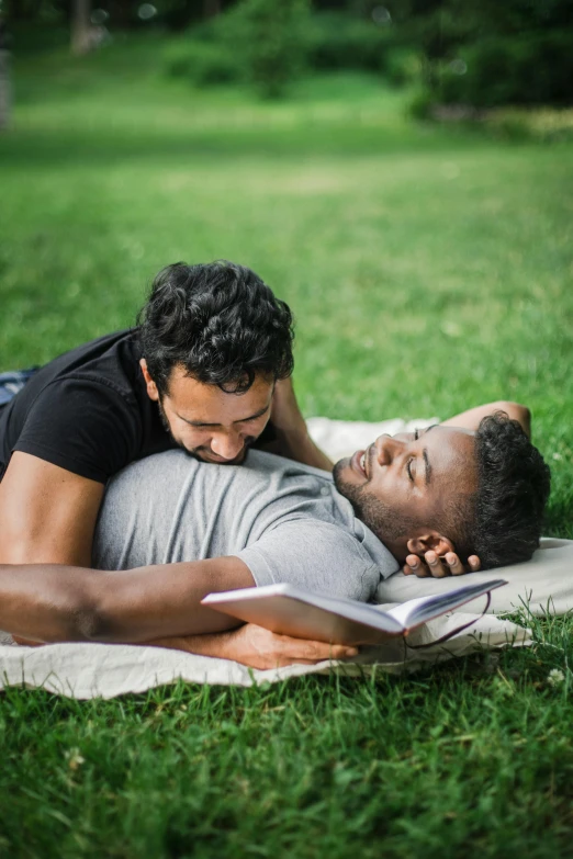 a man laying on a blanket while reading a book, a photo, by Carey Morris, trending on unsplash, romanticism, two men hugging, pride month, in a park, varying ethnicities