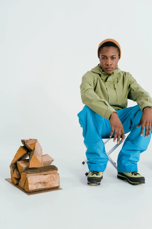 a man sitting on a chair next to a pile of wood, inspired by Afewerk Tekle, visual art, wearing a blue hoodie, androgynous male, climber, green and brown clothes