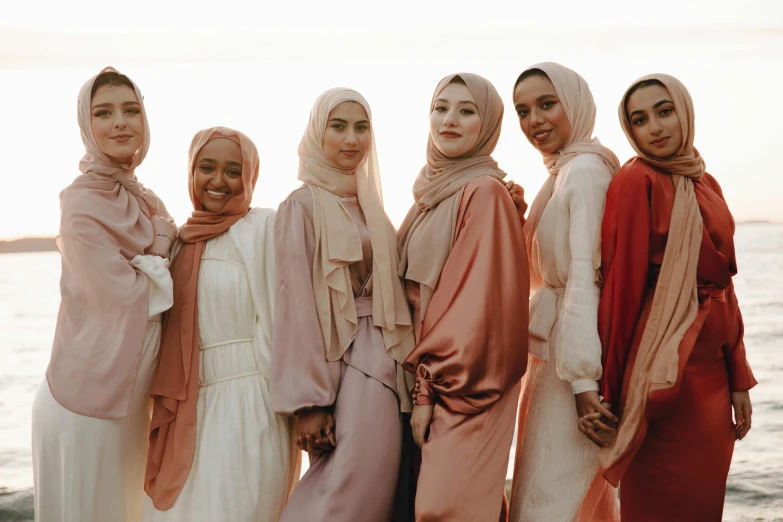 a group of women standing next to each other on a beach, inspired by Maryam Hashemi, trending on pexels, hurufiyya, flowing salmon-colored silk, white hijab, warm skin tone, evening time