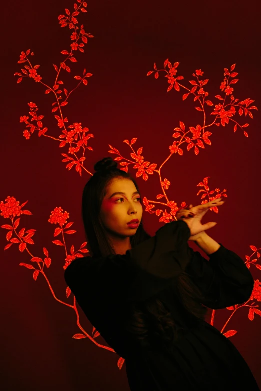 a woman standing in front of a red wall, an album cover, inspired by Gao Xiang, red flowers, lit up in a dark room, pose 4 of 1 6, holding a magic staff