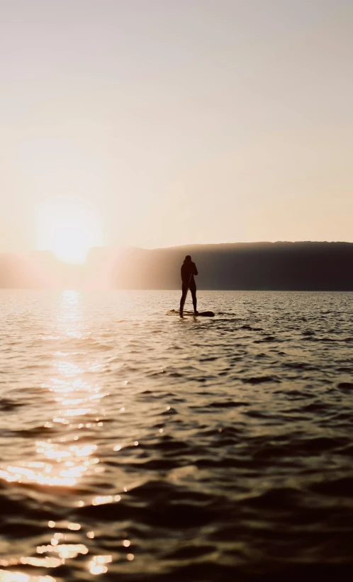 a person standing on a surfboard in the water, by Jessie Algie, on the calm lake surface, late afternoon sun, standup, the dead sea