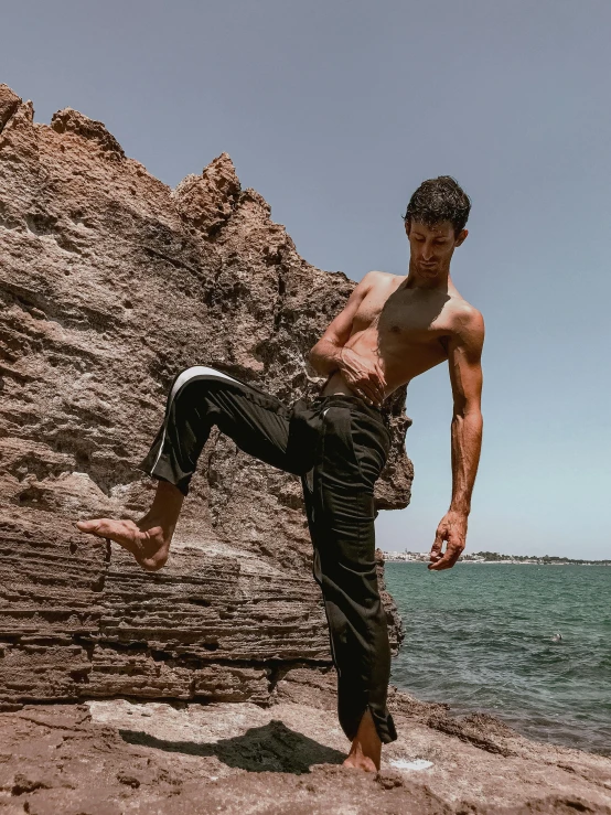 a man standing on top of a rock next to the ocean, inspired by Ma Quan, arabesque, wearing cargo pants, black, posing ready for a fight, profile image