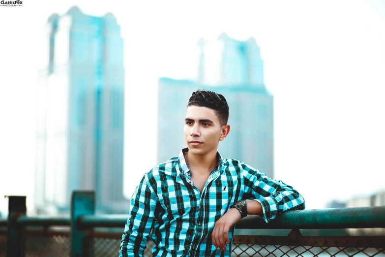 a man leaning on a fence with a city in the background, an album cover, pexels contest winner, realism, middle eastern skin, wearing a light blue shirt, portait photo profile picture, male teenager