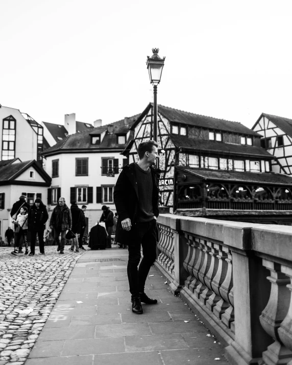 a black and white photo of a man on a bridge, by Tobias Stimmer, town in the background, looking cute, people walking around, the munster in the background