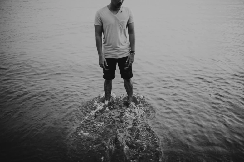 a man standing in the middle of a body of water, a black and white photo, unsplash, wearing shorts and t shirt, on a rock, sad men, lowres