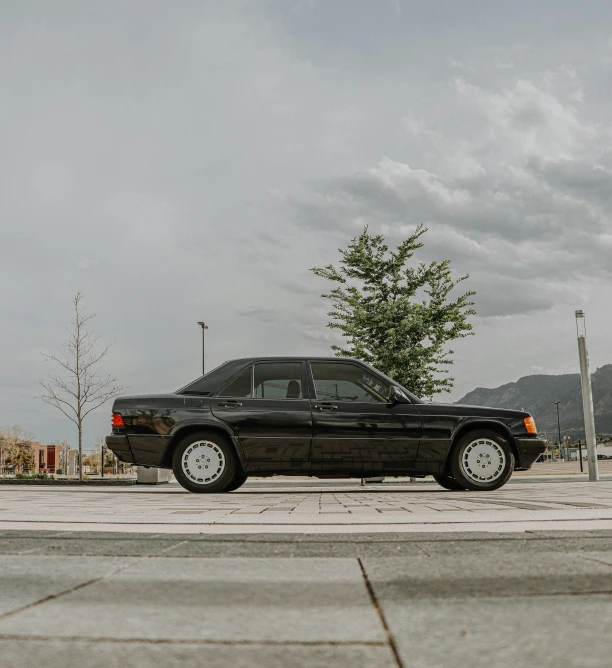 a black car is parked in a parking lot, by Matthias Stom, front view 1 9 9 0, with mountains as background, mercedez benz, panoramic