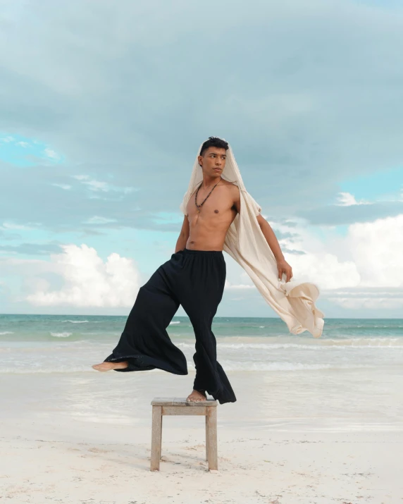 a man standing on a stool on the beach, an album cover, inspired by Byron Galvez, sumatraism, male model, low quality photo, gif, wearing a long flowy fabric