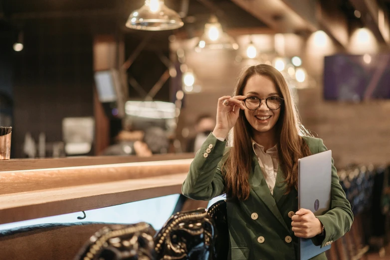 a woman in a green jacket holding a laptop, a portrait, by Emma Andijewska, trending on pexels, standing in a restaurant, girl wearing round glasses, post graduate, happy girl