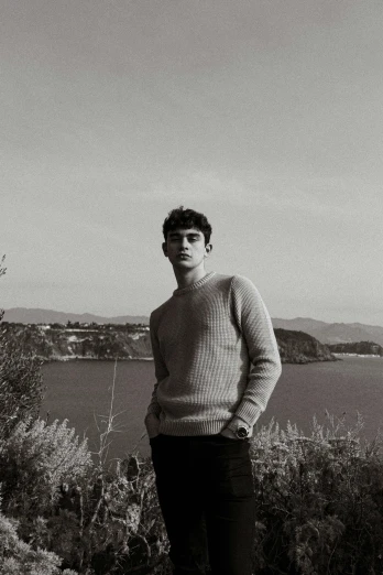 a man standing on top of a hill next to a body of water, a black and white photo, by Alexis Grimou, declan mckenna, he is wearing a brown sweater, portait photo profile picture, robert sheehan