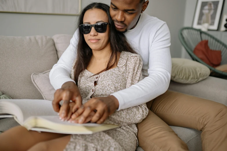 a man and woman sitting on a couch looking at a book, pexels contest winner, wearing shades, pregnant, holding each other hands, diverse