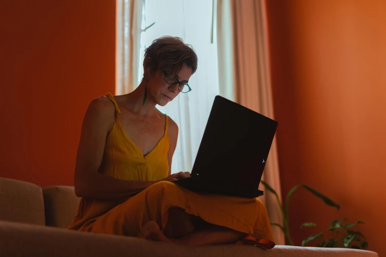 a woman sitting on a couch using a laptop computer, by Carey Morris, pexels, orange yellow ethereal, someone sits in bed, avatar image, serious lighting