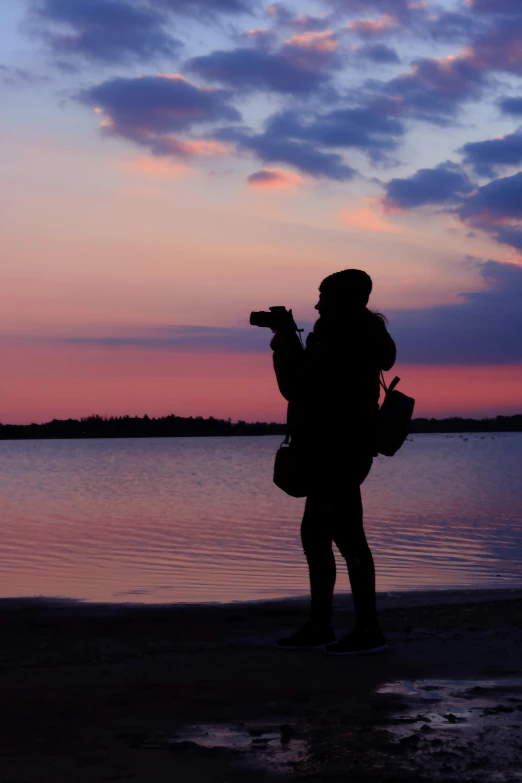 a person standing on a beach holding a camera, pink skies, on a lake, facing the camera, siluette