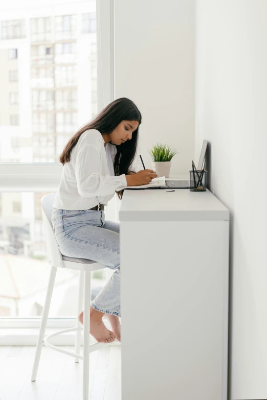 a woman sitting at a desk writing on a piece of paper, by Nicolette Macnamara, clean minimalist design, location in a apartment, profile image, student