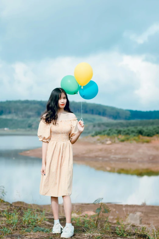 a woman standing in front of a lake holding a bunch of balloons, a colorized photo, by Tan Ting-pho, unsplash, square, 256435456k film, vietnamese woman, earth and pastel colors